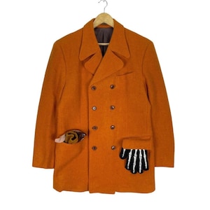 May include: A double-breasted orange wool coat with a brown button closure and two pockets. One pocket has a brown and gold scarf, and the other pocket has a black and white skeleton hand.