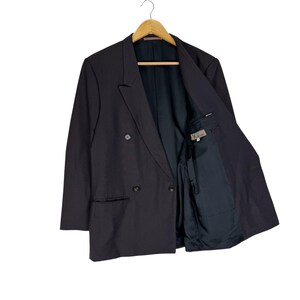 Op de afbeelding: Een zwarte double-breasted blazer met een revers, twee rijen knopen en een zak aan elke kant. De blazer hangt aan een houten hanger. De binnenvoering is zwart.