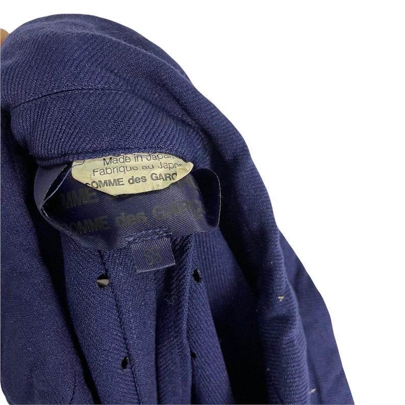 Puede incluir: Una chaqueta azul marino con una etiqueta que dice "Made in Japan, Fabrique au Japon, COMME des GARCONS".