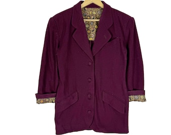 May include: A dark burgundy blazer with a notched lapel, three-button closure, and two front pockets. The cuffs are rolled up, revealing a patterned lining. The blazer is hanging on a wooden hanger.