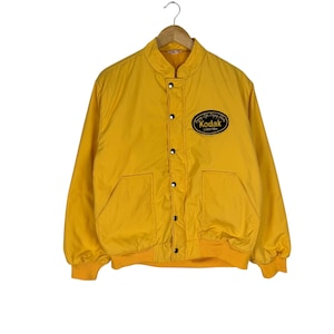 Puede incluir: Chaqueta Kodak amarilla vintage con cierre de botones, dos bolsillos y dobladillo y puños acanalados. La chaqueta presenta un parche ovalado negro con el logotipo de Kodak y el texto "Enjoying Life, Enjoying Film".
