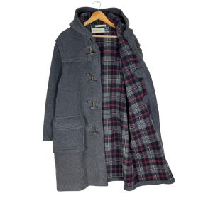 Op de afbeelding: Een grijze duffelcoat met een capuchon en knevelsluitingen. De jas heeft een voering met rood, zwart en wit ruitpatroon. De jas hangt aan een houten kleerhanger.