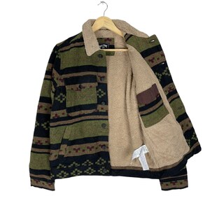 May include: A green, black and brown patterned fleece-lined jacket with a button closure. The jacket has a sherpa lining and a patch pocket on the left side.
