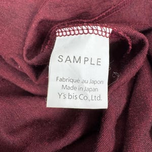 Op de afbeelding: Close-up van een bordeauxrode stof met een wit label. Het label leest "SAMPLE" en bevat tekst in het Frans en Engels: "Fabriqu&eacute; au Japon Made in Japan Y's bis Co.,Ltd."