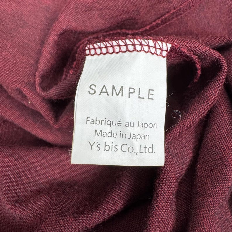 Op de afbeelding: Close-up van een bordeauxrode stof met een wit label. Het label leest "SAMPLE" en bevat tekst in het Frans en Engels: "Fabriqu&eacute; au Japon Made in Japan Y's bis Co.,Ltd."