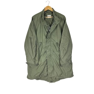 May include: An olive green military-style parka jacket hanging on a wooden hanger. The jacket features a button-up front, a drawstring waist, and multiple pockets. The jacket is made of a durable, possibly water-resistant material.
