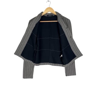 Op de afbeelding: Een gestreepte blazer met een zwarte binnenkant, hangend aan een houten hanger. De blazer heeft verticale strepen in grijstinten en beige. De binnenkant is zwart met een rasterpatroon.