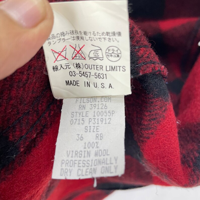 Op de afbeelding: Een rood-zwart geruit overhemd met een wit label. Het label heeft de tekst "FILSON.COM", "RN 39126", "STYLE 10055P", "0715 P31912", "SIZE 36 RB", "100% VIRGIN WOOL", "PROFESSIONALLY DRY CLEAN ONLY".