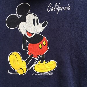 K&ouml;nnte beinhalten: Ein marineblaues Sweatshirt mit einem Cartoon-Bild von Mickey Mouse, der rote Shorts und gelbe Schuhe tr&auml;gt. Der Text "California" ist in Wei&szlig; &uuml;ber Mickey Mouse gedruckt und der Text "&copy; THE WALT DISNEY COMPANY BY VELVA SHEEN" ist in Wei&szlig; unter Mickey Mouse gedruckt.