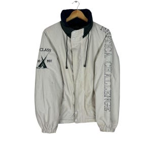 Puede incluir: Una chaqueta beige con forro de polar negro y verde. La chaqueta tiene cierre de cremallera y capucha con cordón. La manga izquierda tiene un logotipo que dice "CLASS 1937" y un gráfico de dos veleros. La manga derecha tiene el texto "NAUTICA CHALLENGE" impreso verticalmente.