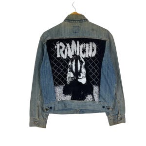 May include: A light blue denim jacket with a black graphic on the back. The graphic features the word "RANCID" above a black and white image of a figure behind a chain-link fence. The jacket has a button-up front and two chest pockets.