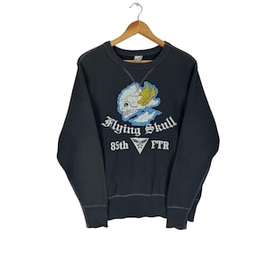 May include: A dark gray sweatshirt with a graphic featuring a skull with wings, clouds, and the text "Flying Skull" and "85th FTR" in white. The design is in blue, yellow, and white. The sweatshirt is hanging on a wooden hanger.