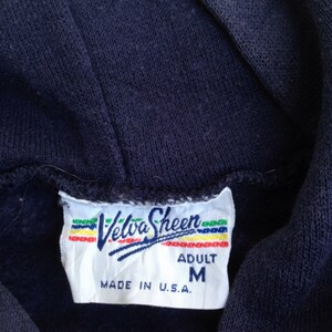 K&ouml;nnte beinhalten: Ein wei&szlig;es Etikett mit einem blauen und roten Logo, auf dem "Velva Sheen" steht, sowie dem Text "Adult M Made in U.S.A."