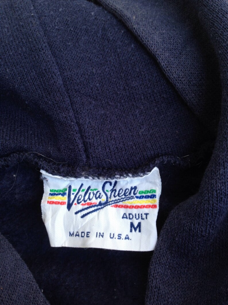 K&ouml;nnte beinhalten: Ein wei&szlig;es Etikett mit einem blauen und roten Logo, auf dem "Velva Sheen" steht, sowie dem Text "Adult M Made in U.S.A."