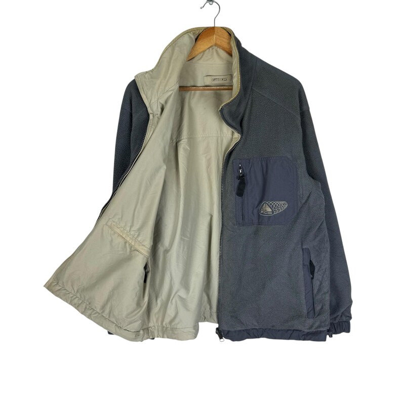 Puede incluir: Chaqueta polar gris y beige con cremallera y cuello alto. La chaqueta tiene un bolsillo azul marino con un logotipo y una cremallera negra. La chaqueta est&aacute; colgada de una percha de madera.