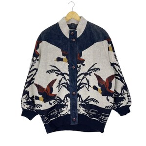 Könnte beinhalten: Ein cremefarbener Strickcardigan mit einem blauen Wildlederjoch und einem Muster aus braunen und roten Enten, die über einem weißen Hintergrund fliegen. Der Cardigan hat einen Knopfschließe vorne und lange Ärmel.