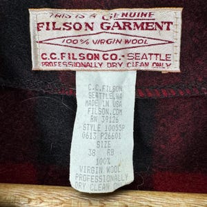 Puede incluir: Primer plano de una etiqueta de prenda Filson. La etiqueta dice "THIS IS A GENUINE FILSON GARMENT" y "100% VIRGIN WOOL". Debajo, se lee "C.C. FILSON CO. SEATTLE" y "PROFESSIONALLY DRY CLEAN ONLY". Una etiqueta m&aacute;s peque&ntilde;a enumera detalles como la talla 38 RB y "100% VIRGIN WOOL".