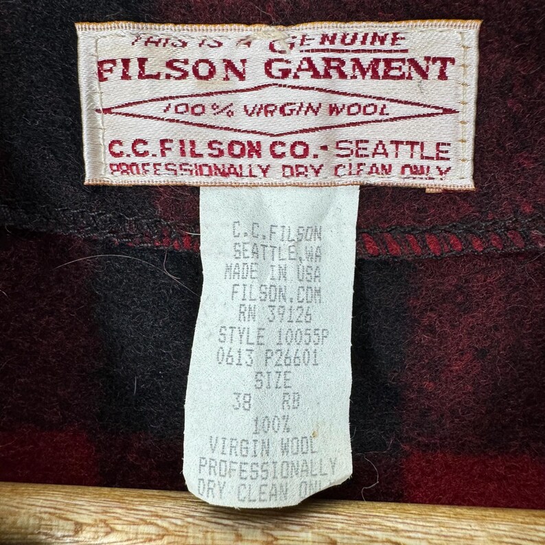 Puede incluir: Primer plano de una etiqueta de prenda Filson. La etiqueta dice "THIS IS A GENUINE FILSON GARMENT" y "100% VIRGIN WOOL". Debajo, se lee "C.C. FILSON CO. SEATTLE" y "PROFESSIONALLY DRY CLEAN ONLY". Una etiqueta m&aacute;s peque&ntilde;a enumera detalles como la talla 38 RB y "100% VIRGIN WOOL".