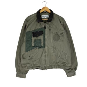 May include: A green utility jacket with a black collar and multiple pockets. The jacket has a zipper closure and a patch with the word "Winston" on the left chest.
