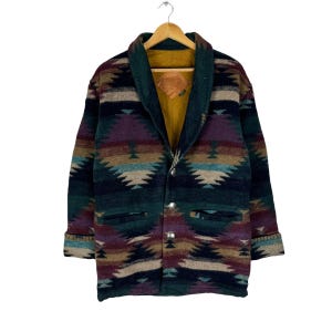 Könnte beinhalten: Eine langärmelige Jacke mit Schalkragen und Knopfleiste. Die Jacke ist in Dunkelgrün, Braun, Lila und Blau gemustert und hat zwei Taschen auf der Vorderseite und einen rautenförmigen Lederfleck auf der Innenseite.