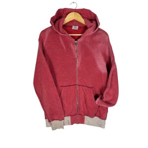 May include: A red zip-up hoodie with a hood and front pockets, hanging on a wooden hanger. The bottom and cuffs are light grey. The hoodie is made of a soft, comfortable material. A casual garment.