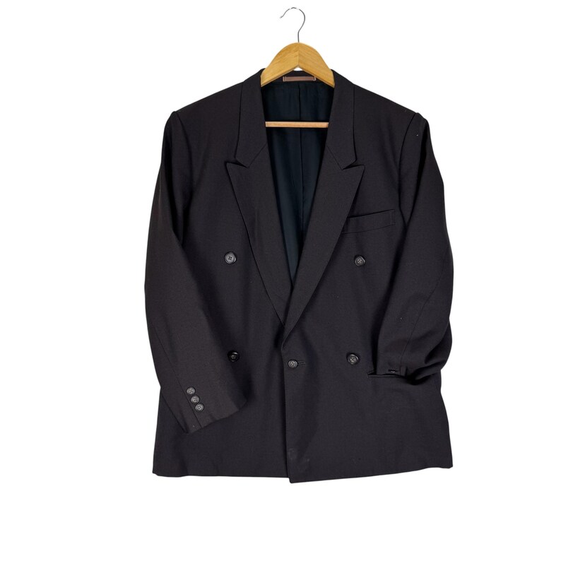 Op de afbeelding: Een zwarte double-breasted blazer hangt aan een houten hanger. De blazer heeft een revers, twee rijen knopen en drie knopen op elke manchet. De jas is gemaakt van een effen zwarte stof.