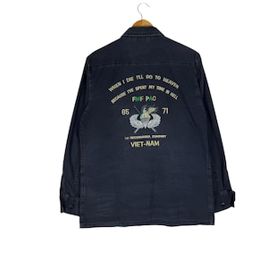 Könnte beinhalten: Dunkelmarineblaues langärmliges Button-up-Hemd. Auf der Rückseite befindet sich ein gestickter Text: "WHEN I DIE I'LL GO TO HEAVEN BECAUSE I'VE SPENT MY TIME IN HELL" mit "FMF PAC", "65", "71" und "1st RECONNAISSA COMPANY VIET-NAM".