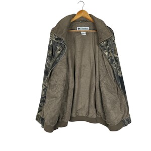 May include: A Columbia camouflage jacket with a brown interior and a zip-up front. The jacket is hanging on a wooden hanger. The jacket has a collar and a logo on the chest.