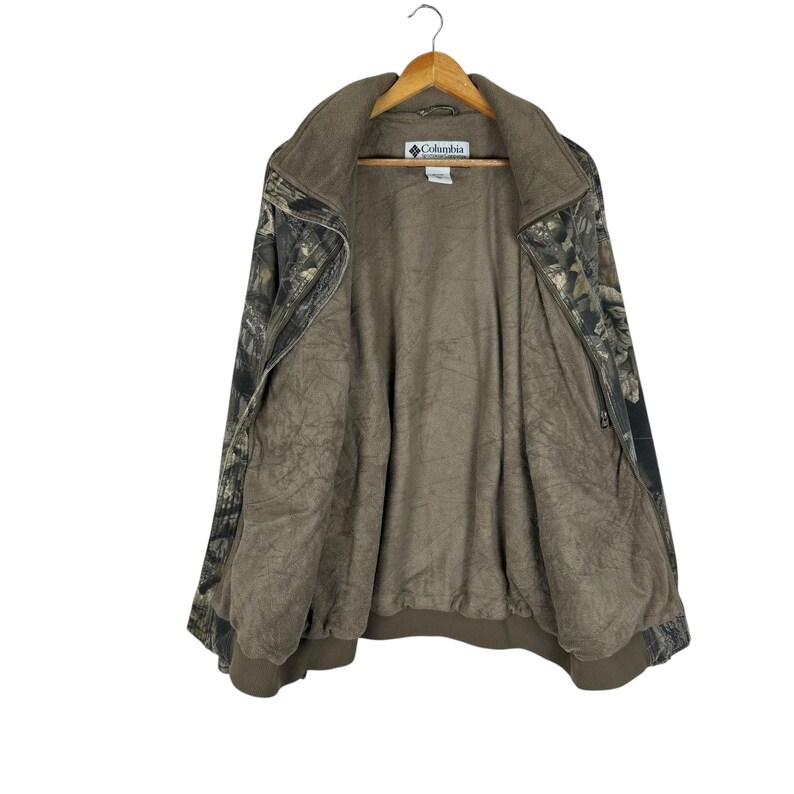 May include: A Columbia camouflage jacket with a brown interior and a zip-up front. The jacket is hanging on a wooden hanger. The jacket has a collar and a logo on the chest.