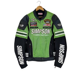 May include: A green and black Simpson racing jacket with white accents. The jacket features the Simpson logo across the chest and sleeves, along with various patches. The jacket is hanging on a wooden hanger.