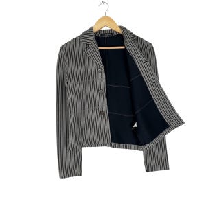 Op de afbeelding: Een gestreepte blazer in grijstinten en zwart hangt aan een houten hanger. De blazer heeft een klassiek ontwerp met een reverskraag, knoopsluiting en lange mouwen. De binnenvoering is zwart.