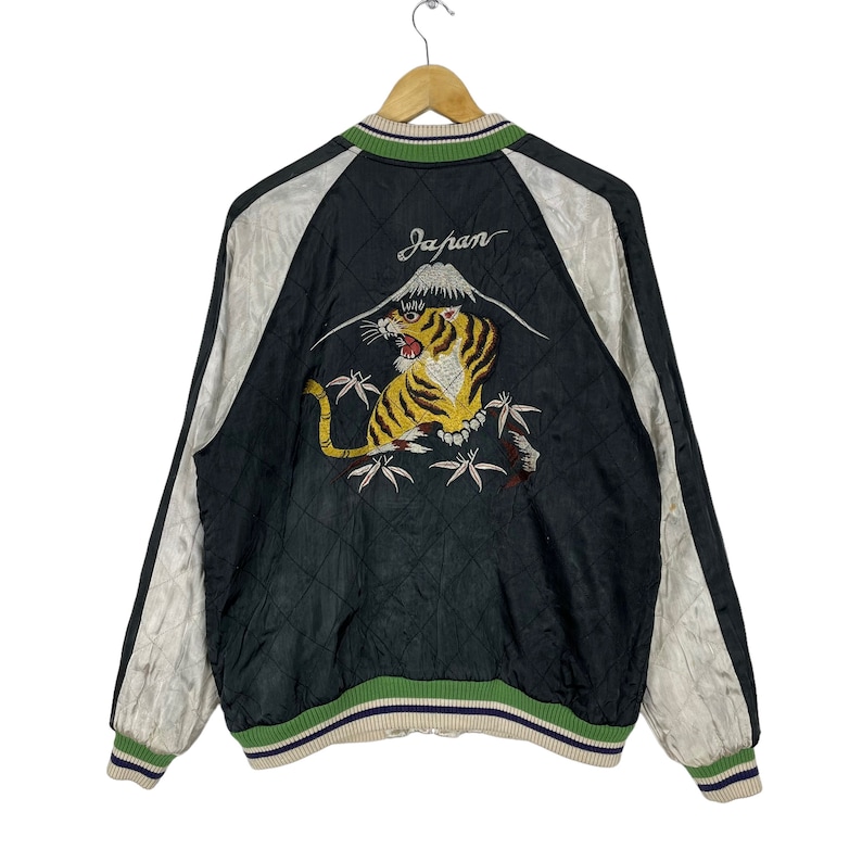 May include: Black satin bomber jacket with a white tiger embroidered on the back. The tiger is surrounded by bamboo and a mountain with the word "Japan" above it. The jacket has green and white striped ribbing at the collar, cuffs, and hem.