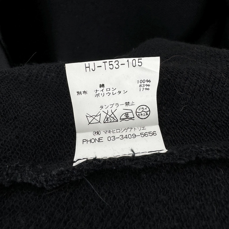 May include: A white tag with black text and symbols. The tag has the text "HJ-T53-105" at the top. The tag also has the text "綿 100% ポリウレタン 83% 17%" and "タンブラー禁止" in Japanese. The tag also has a phone number: "PHONE 03-3409-5656".