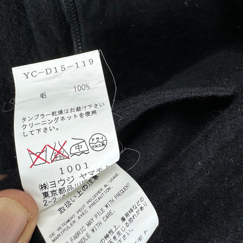 K&ouml;nnte beinhalten: Ein schwarzes Pflegeetikett f&uuml;r Kleidung mit wei&szlig;em Text. Das Etikett lautet "YC-D15-119", "100%", "毛", "タンブラー乾燥はお避け下さい", "クリーニングネットを使用して下さい", "1001", "(株)ヨウジ", "東京都品川", "2-2-", "取扱い上の注意", "ドライ", "セキス", "CE VETEMENT PEUT PELUCHER AL USAGE", "MANIPULER AVEC PRECAUTION", "FABRIC MAY PILE WITH FREQUENT", "HANDLE WITH CARE", "の特性上、着用時などの", "下さい", "みを生じる恐れがあり", "モト".