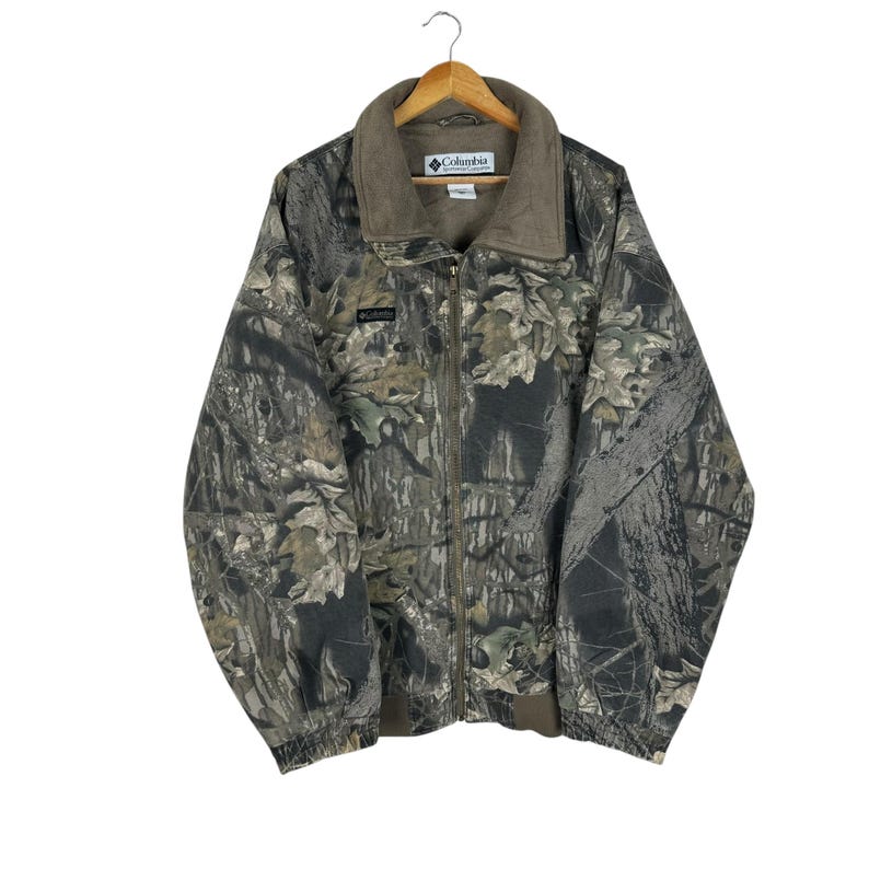 May include: A camouflage Columbia jacket with a full zip and a brown fleece collar. The jacket has a woodland pattern in green, brown, and grey. The Columbia logo is visible on the chest and collar.