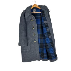 May include: A gray wool duffel coat with a hood and wooden toggles. The coat's interior features a blue and black plaid pattern. The coat is hanging on a wooden hanger.
