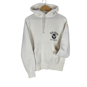 May include: Cream-colored hoodie with a drawstring hood and a kangaroo pocket. The front features a black and white emblem with the text "U.S. ARMY AIR FORCES". The sweatshirt is hanging on a wooden hanger.