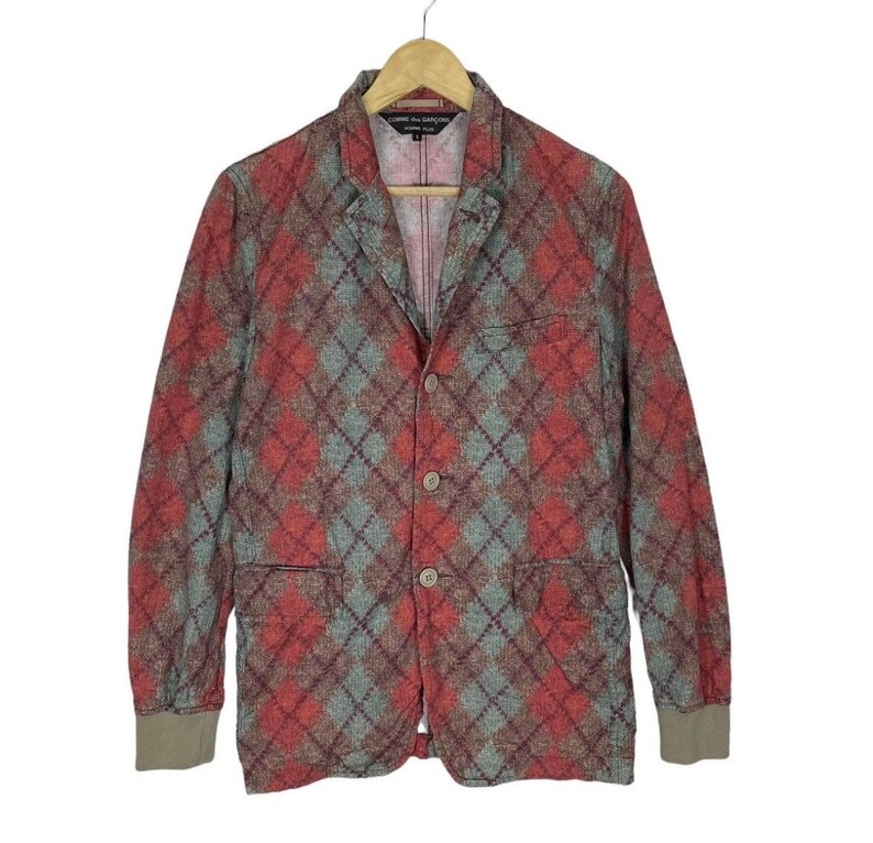 May include: A red, grey, and brown argyle patterned blazer with three buttons and two front pockets. The blazer is hanging on a wooden hanger. The cuffs are a solid brown colour.