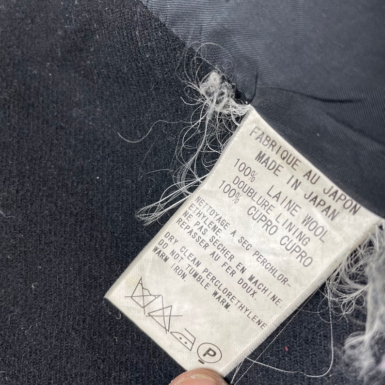 May include: White tag with black text that reads "Fabrique au Japon Made in Japan 100% Laine Wool Doublure Lining 100% Cupro Cupro" and washing instructions. The tag is attached to a black fabric.