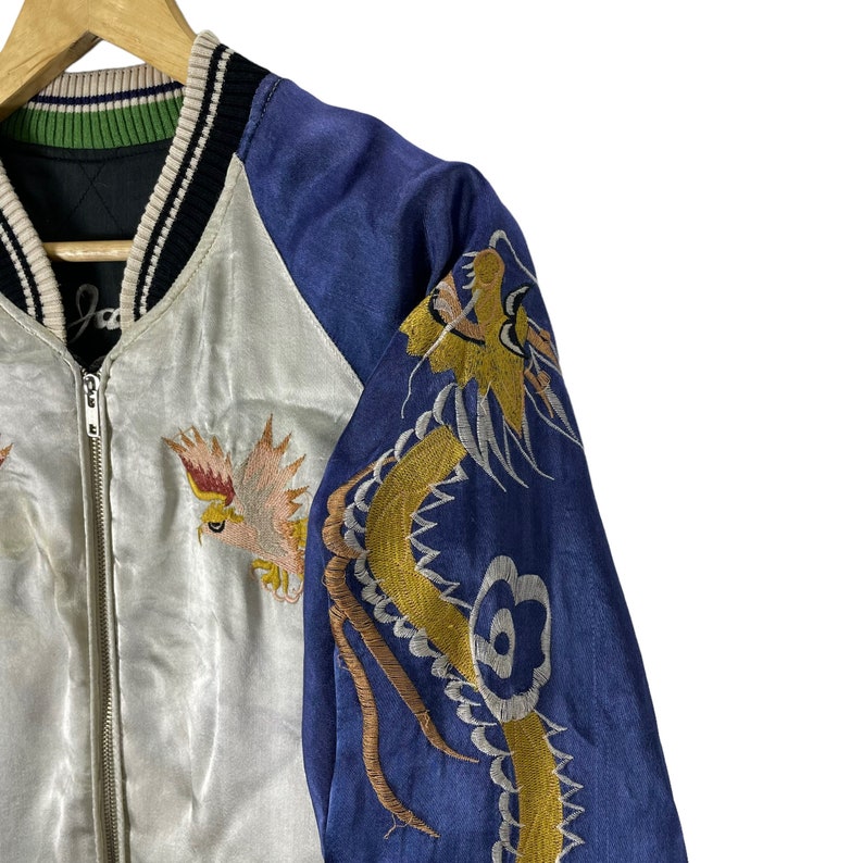 May include: A white and blue satin bomber jacket with embroidered dragons and birds. The jacket has a black and white ribbed collar and a zipper closure.
