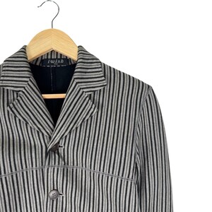 Op de afbeelding: Een grijs en zwart gestreepte blazer met een reverskraag en twee bruine knopen. De blazer hangt aan een houten kleerhanger. Het label luidt "i wish".