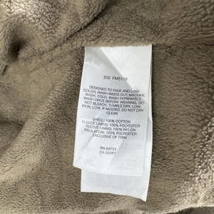 May include: A close-up of a white label with black text, attached to a beige fabric. The label provides washing instructions and fabric details: 100% cotton shell, 100% polyester fleece lining, 100% nylon sleeve lining, and 100% polyester insulation.