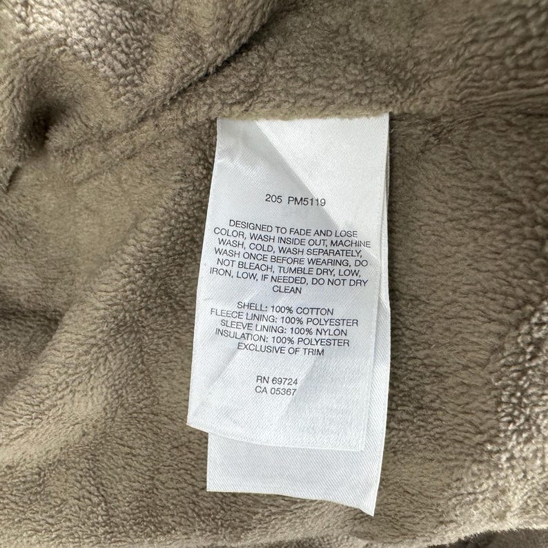 May include: A close-up of a white label with black text, attached to a beige fabric. The label provides washing instructions and fabric details: 100% cotton shell, 100% polyester fleece lining, 100% nylon sleeve lining, and 100% polyester insulation.