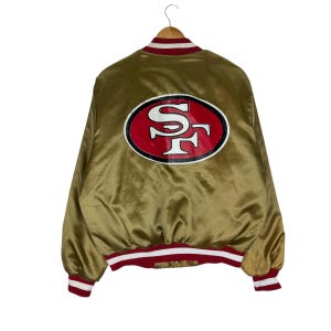 May include: A gold satin jacket with a red, white, and gold logo on the back. The jacket has red and white striped trim on the collar, cuffs, and waist. The logo features the letters "SF" in white on a red oval.