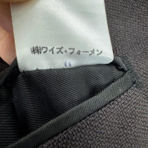 Op de afbeelding: Close-up van een wit label met Japanse tekst, bevestigd aan een donkerbruine stof. Het label is rechthoekig met afgeronde hoeken. De stof is een geweven textuur, waarschijnlijk onderdeel van een kledingstuk.