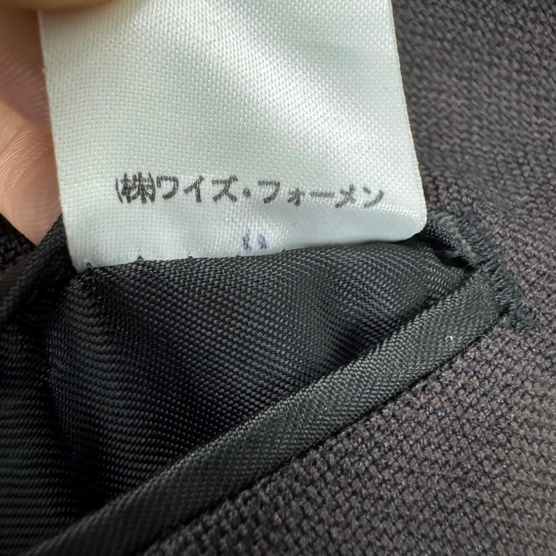 Op de afbeelding: Close-up van een wit label met Japanse tekst, bevestigd aan een donkerbruine stof. Het label is rechthoekig met afgeronde hoeken. De stof is een geweven textuur, waarschijnlijk onderdeel van een kledingstuk.