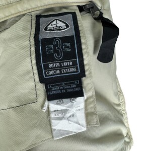 Puede incluir: Primer plano de una prenda beige con una etiqueta negra. La etiqueta presenta el texto "ACG", "3", "OUTER LAYER" y "COUCHE EXTERNE". Debajo de la etiqueta hay una etiqueta m&aacute;s peque&ntilde;a con el texto "MEN'S". Una cremallera y una correa negras son visibles.