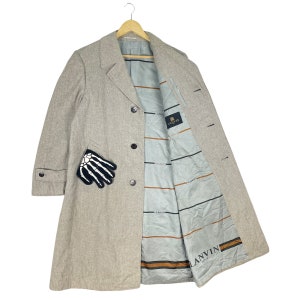 Puede incluir: Abrigo beige claro con un parche de mano de esqueleto en blanco y negro. El abrigo tiene cierre de botones y forro interior azul claro con el logo "LANVIN PARIS". Cuelga de una percha de madera.