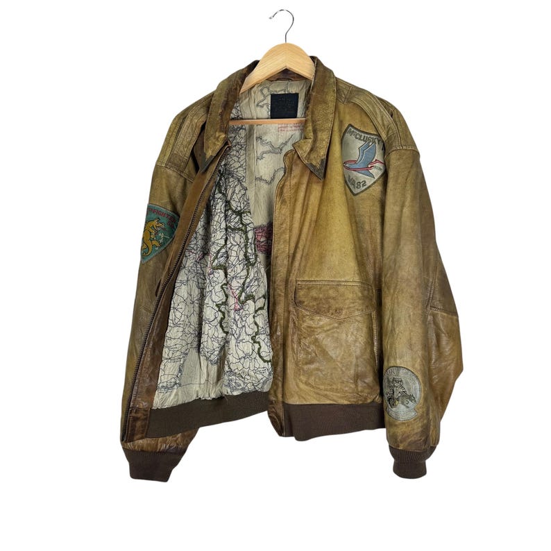 May include: A vintage brown leather bomber jacket with patches and a map-printed lining. The jacket has a worn, distressed appearance, with a zipper closure and ribbed cuffs and hem. Patches include an emblem with a bird and the text "APOLLO 82".