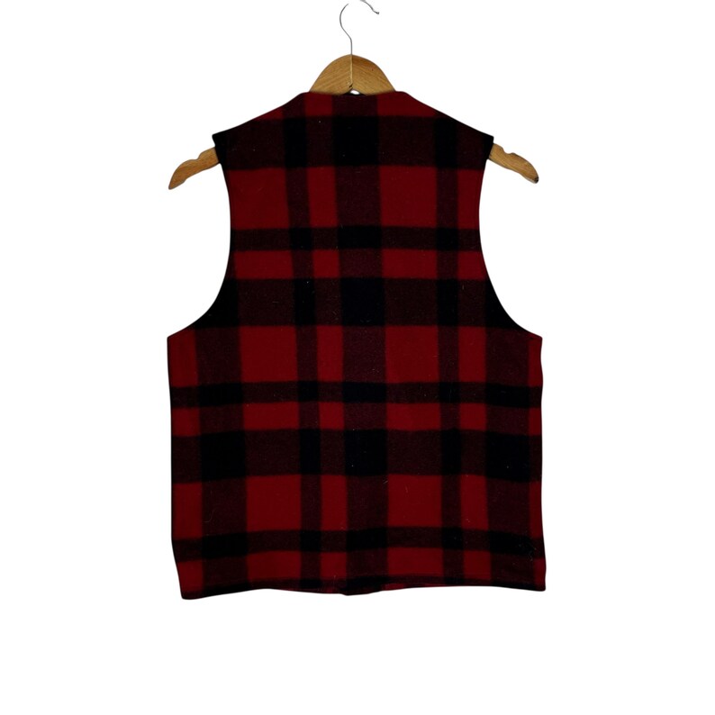 Puede incluir: Un chaleco a cuadros rojos y negros colgado de una percha de madera. El chaleco tiene un estilo cl&aacute;sico de le&ntilde;ador con una parte delantera abotonada y cuello en V. La tela parece ser lana gruesa o una mezcla de lana.